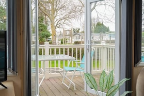 A view through a glass door to a wooden deck with a table and chairs, a white railing, and a green lawn.