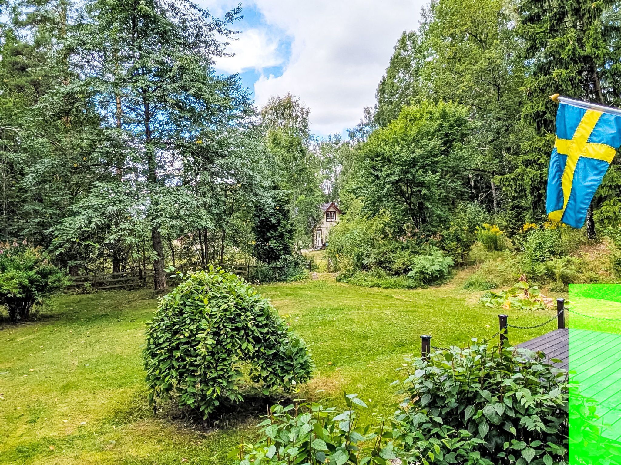 A well-maintained garden with a variety of trees and shrubs, a small house in the background, and a Swedish flag.