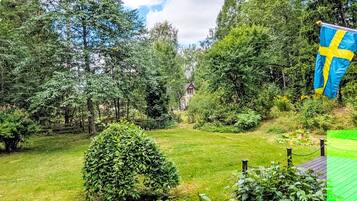 A well-maintained garden with a variety of trees and shrubs, a small house in the background, and a Swedish flag.