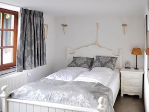 A bedroom with a white bed, grey patterned curtains, a white bedside table with a lamp, and a wall-mounted clock. - Luxury Ardennes Villa With Pool and Sauna (Stoumont)