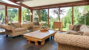 A spacious room with wicker furniture, a wooden coffee table, and large windows offering a view of greenery. - Luxury Ardennes Villa With Pool and Sauna (Stoumont)
