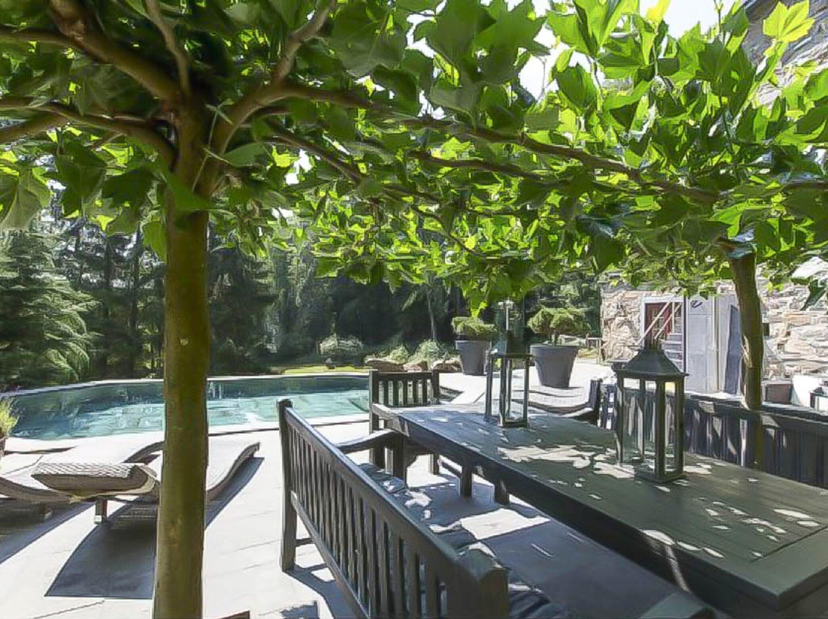 A poolside area with a table and chairs, surrounded by greenery and a stone wall.