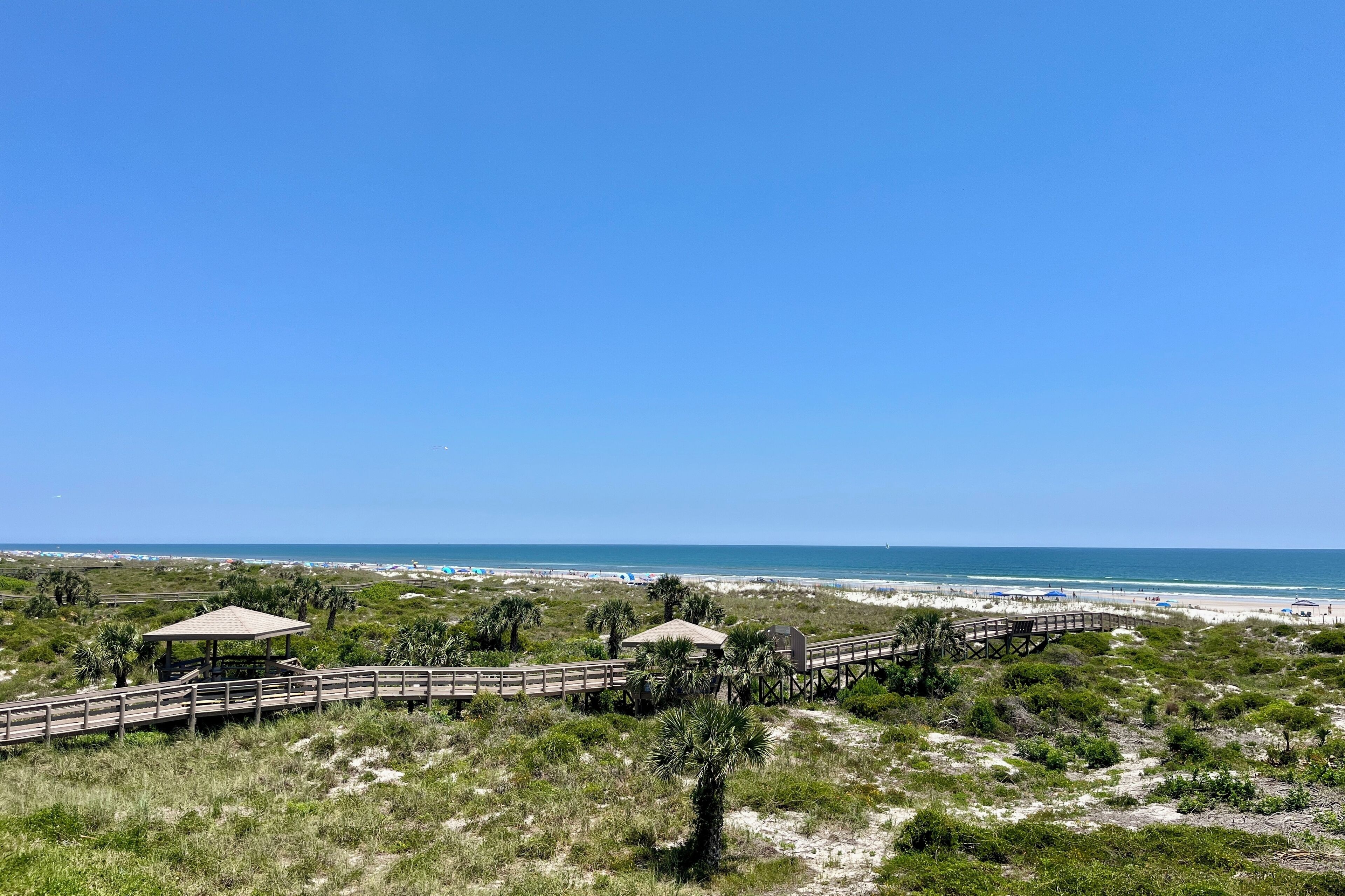 A beach view with a wooden boardwalk, a gazebo, and a beach area with umbrellas.