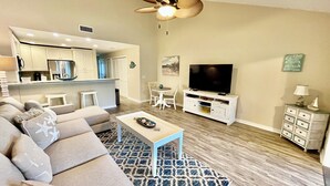 A living room with a sofa, coffee table, and TV. There is a kitchen in the background with cabinets and a refrigerator. - Modern Gold-rated beach chic condo with views of bubbling lagoon! (Saint Augustine)