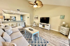 A living room with a sofa, coffee table, TV, and a ceiling fan. - Modern Gold-rated beach chic condo with views of bubbling lagoon! (Saint Augustine)