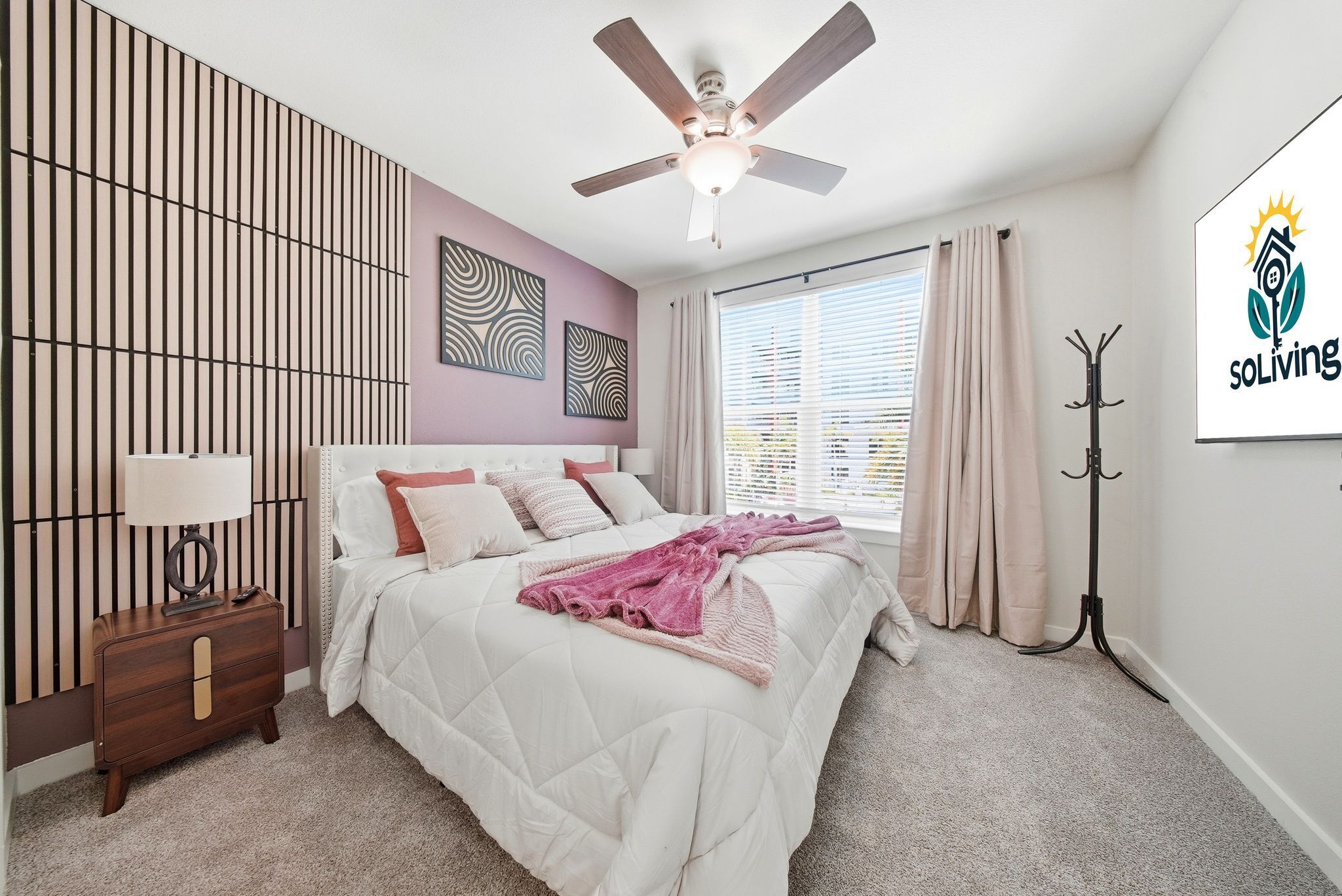 Une chambre à coucher comprenant un lit, un ventilateur de plafond, une table de chevet, un porte-manteau et une fenêtre avec des rideaux.