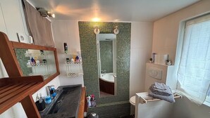 Une salle de bain avec une baignoire, un lavabo, des toilettes et un miroir. On y trouve des étagères avec des articles de toilette et une fenêtre avec des rideaux.