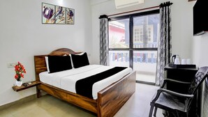 A hotel room with a wooden bed, a desk with a chair, a window with curtains, and a wall-mounted air conditioner.