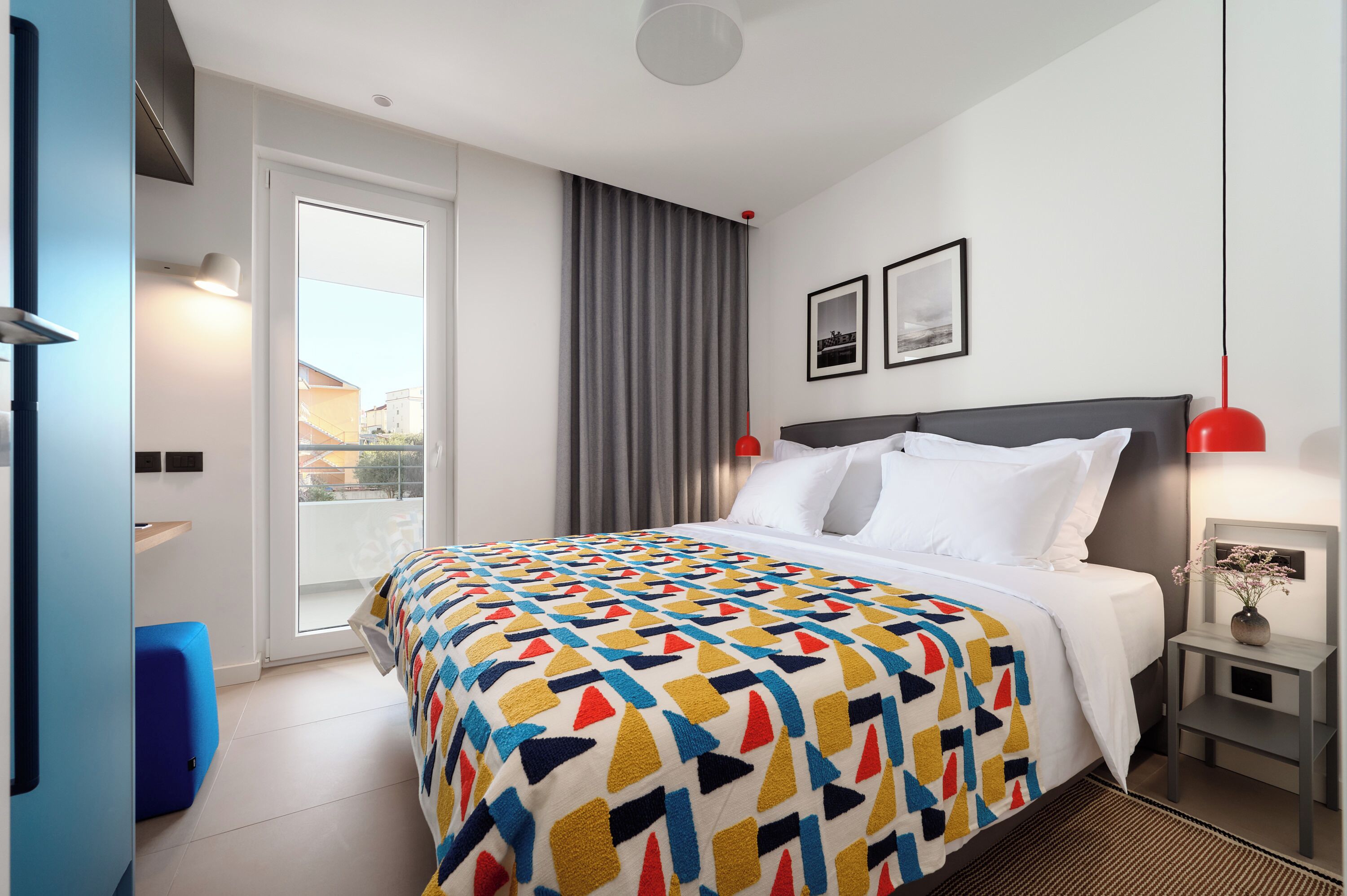 A hotel room with a bed, a window with curtains, a wall-mounted lamp, and a small table with a vase.