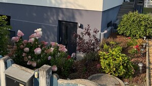 A modern two-story building with a grey and white facade, a small front garden with pink flowers, and a wooden fence with a sign. - Cozy quiet apartment in Berlin with terrace, free parking (Berlin)