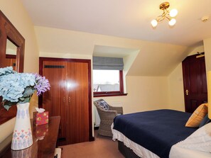 A bedroom with a wooden wardrobe, a bedside table with a vase and a tissue box, a wicker chair, a bed with a blue blanket, and a window with a view of the outside. - Dunhallin House (Inverness)