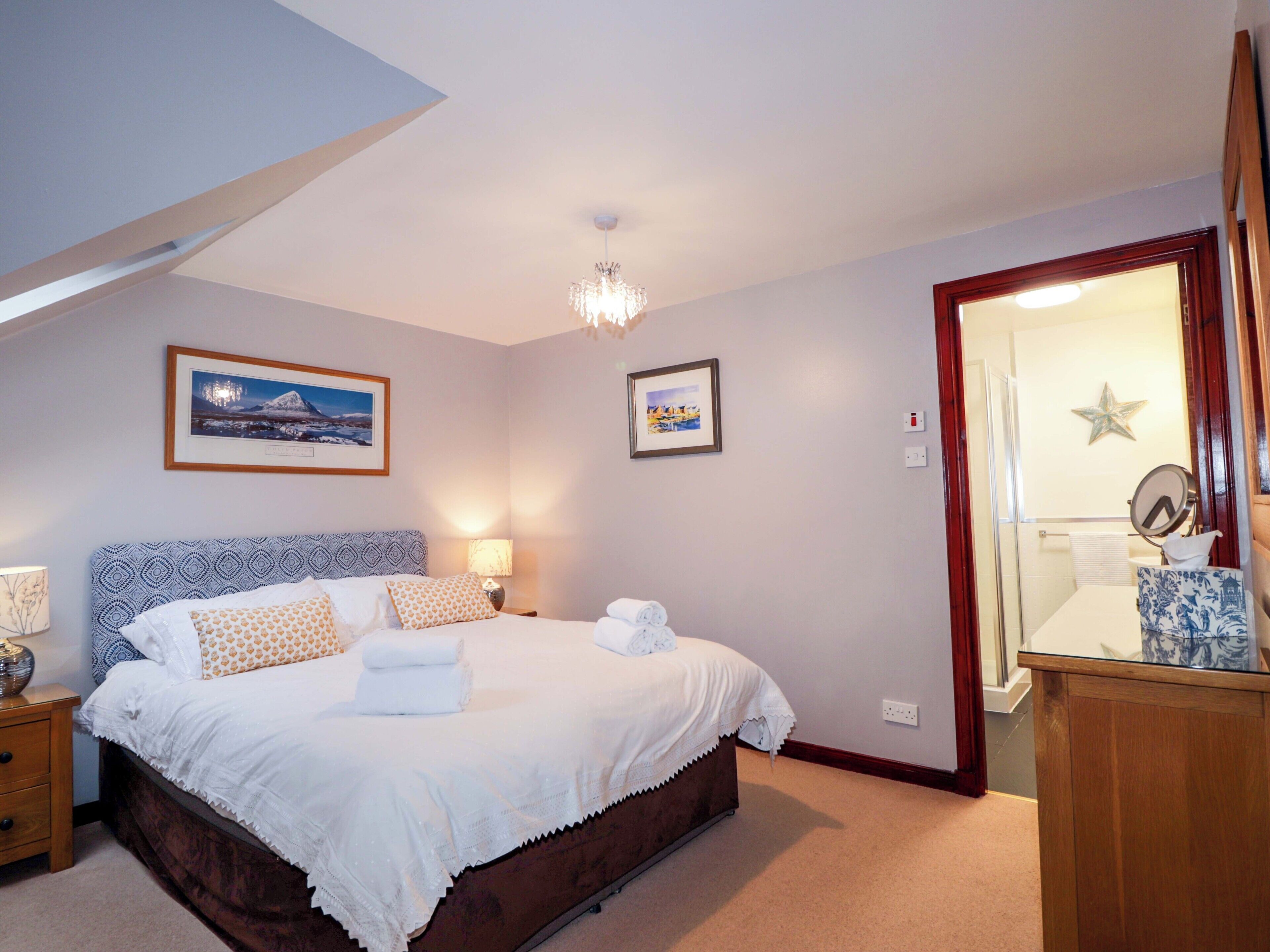 A well-lit bedroom with a large bed, bedside lamps, a chandelier, framed pictures on the wall, and a wooden door leading to a bathroom.