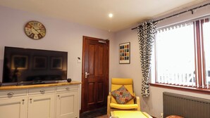 A living room with a flat-screen TV, a wooden door, a yellow armchair, a patterned curtain, and a bench. - Dunhallin House (Inverness)