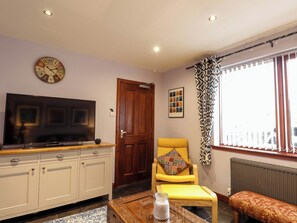 A living room with a flat-screen TV, a wooden door, a yellow armchair, a patterned curtain, and a bench. - Dunhallin House (Inverness)