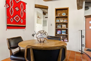 A dining area with a round wooden table, two chairs, a decorative metal sphere, a red patterned cloth, and a bookshelf with books and decorative items.
