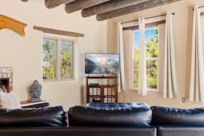 A living room with a leather sofa, a television on a wooden stand, and two windows with curtains.