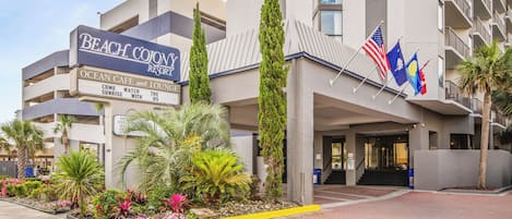 A hotel entrance with a sign for Beach Colony Resort, featuring a landscaped garden with tropical plants and a paved walkway.