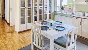 A dining area with a white table set for four, surrounded by white chairs. A kitchen with white cabinets and a window is visible in the background. - Tallkotten by Interhome (Storebro)