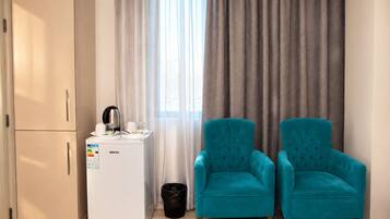 Une chambre d’hôtel équipée d’un mini-bar, de deux fauteuils turquoise, d’un lit, d’une armoire blanche et d’une fenêtre avec des rideaux.