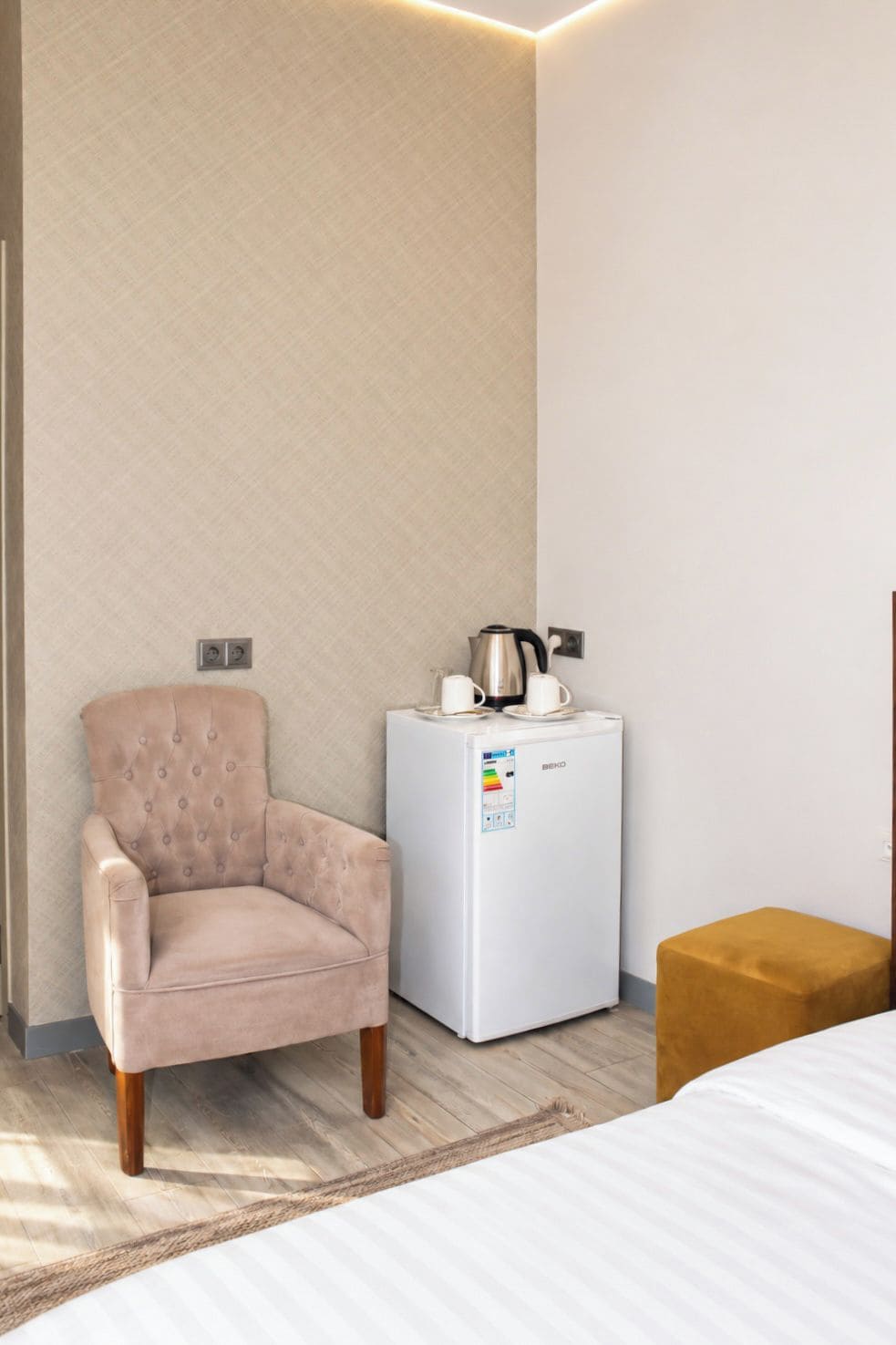 Una habitación con cama, una silla, un refrigerador blanco pequeño y una mesita de noche de madera.