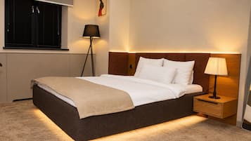 A hotel room with a bed, bedside table, lamp, and a window with blinds.