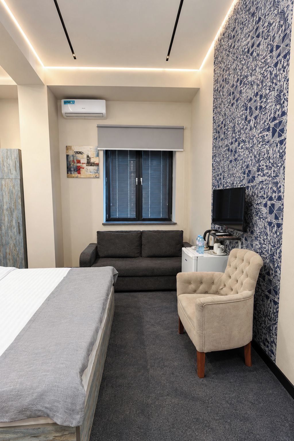 A hotel room with a bed, a sofa, a chair, a TV, and a small table with a kettle and a bottle.