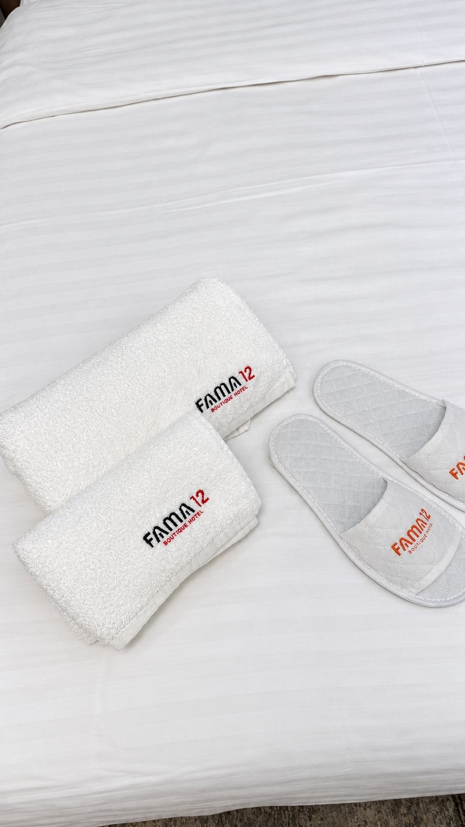 Un par de pantuflas blancas y una toalla blanca con el logo de 'FAMA 12 Boutique Hotel'.
