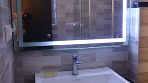A bathroom with a white sink, a mirror with a light, and a tiled wall. - District Gravity (Shamirpet)