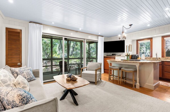 A modern living room with a sofa, a coffee table, and a kitchen area with a large window, a TV, and a bar. - Beach access, close to West Beach conference (Kiawah Island)