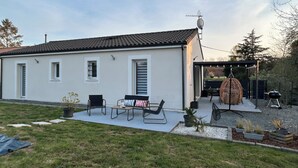 Una casa de un piso con techo de tejas, un patio con banco y mesa, una silla colgante y una parrilla.