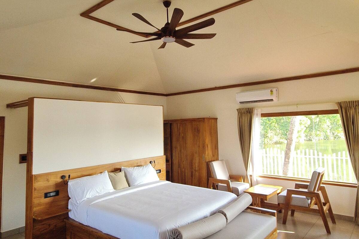 Un dormitorio con cama de madera, ventilador de techo, ventana con cortinas y vista a zonas verdes.
