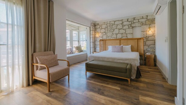 A bedroom with a stone wall, a bed, a chair, and a window with a view. - Fiora Alacati Hotel (Çeşme)
