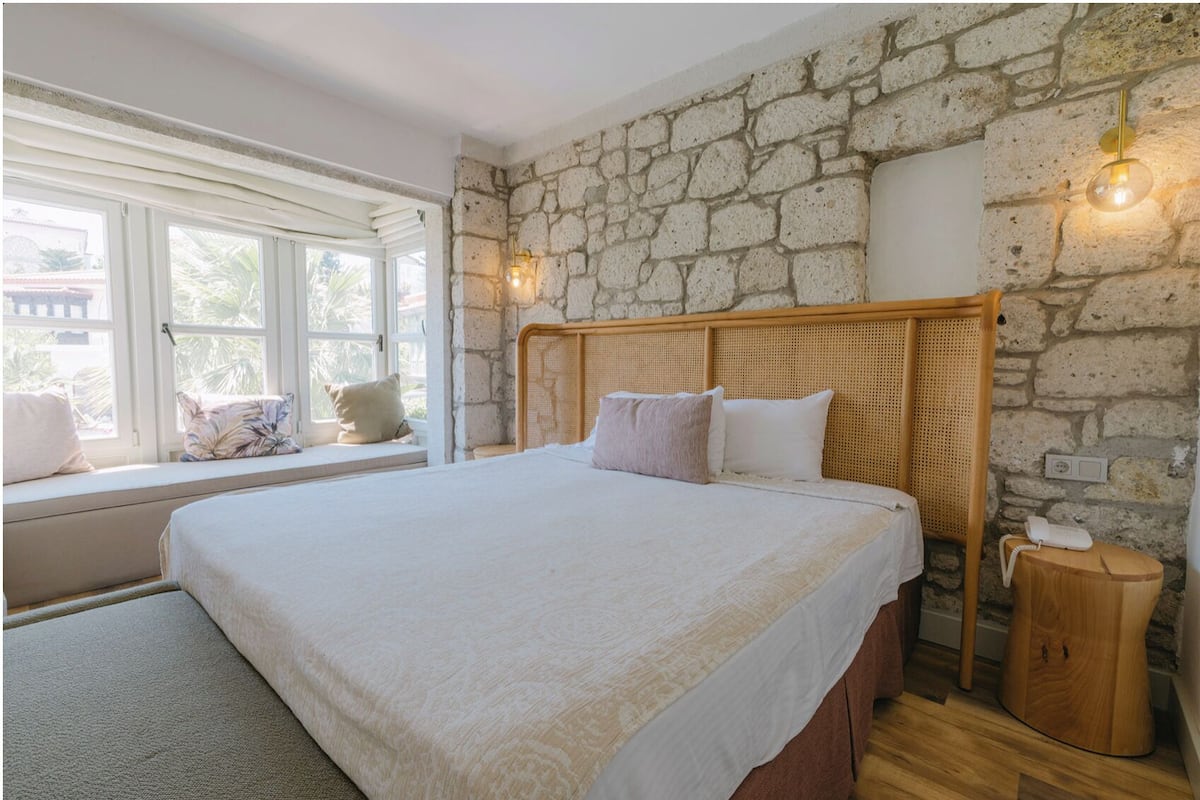 A bedroom with a stone wall, a large bed, a bedside table, a nightstand, and a window with a view.