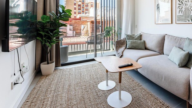 Uma sala de estar moderna com um sofá, uma televisão, uma mesinha, um lustre e uma varanda com vista para edifícios.