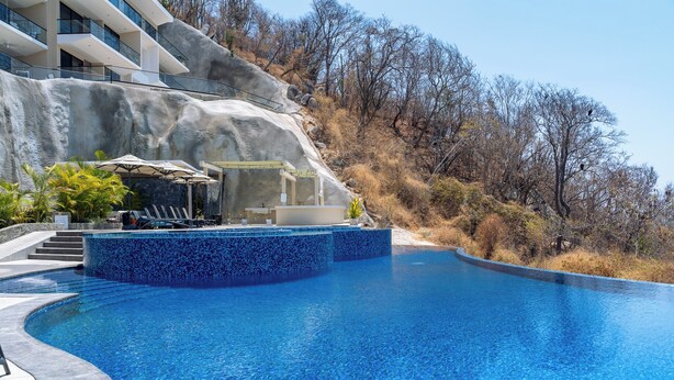 Una piscina con agua cristalina azul, un muro de piedra y un edificio con balcones al fondo.