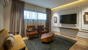 A hotel room with a sofa, two armchairs, a coffee table, a flat-screen TV mounted on the wall, and a large window with curtains. - Joannes (Abidjan)