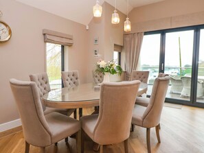 A dining room with a round glass table, six upholstered chairs, a vase with flowers, and a view through large windows. - Swing Bridge Cottage (Driffield)