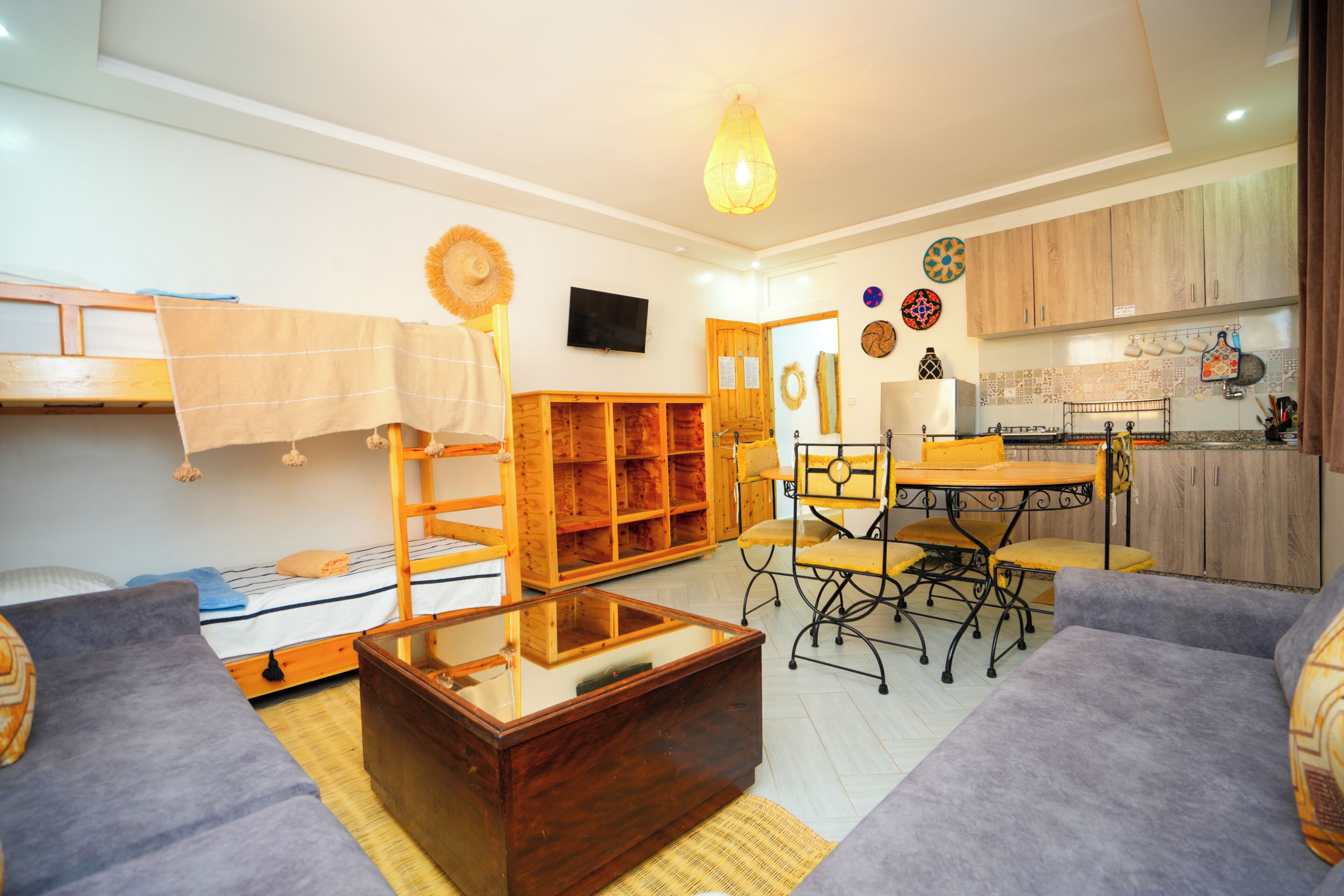 Un espacio habitable pequeño con una cama litera, un cofre de madera, una mesa de comedor con sillas y una zona de cocina.