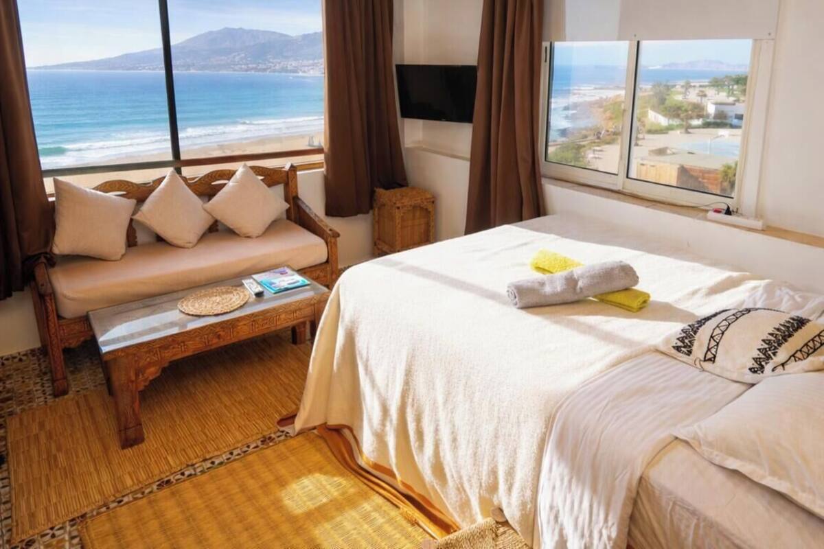 A bedroom with a bed, a bench, a view of the ocean, and a window.