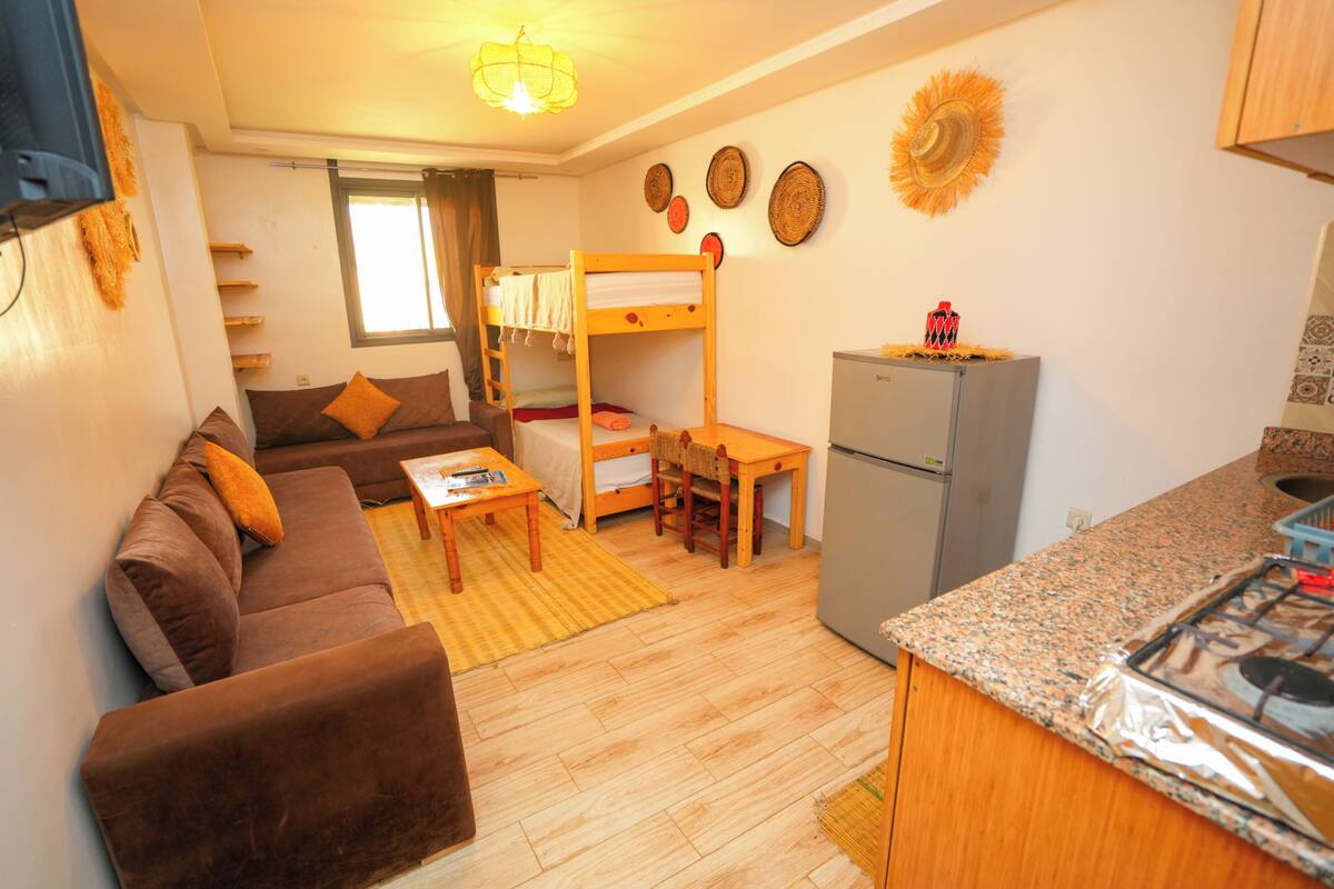 A compact living space with a sofa, bunk beds, a small table, a refrigerator, and a kitchen area with a stove.