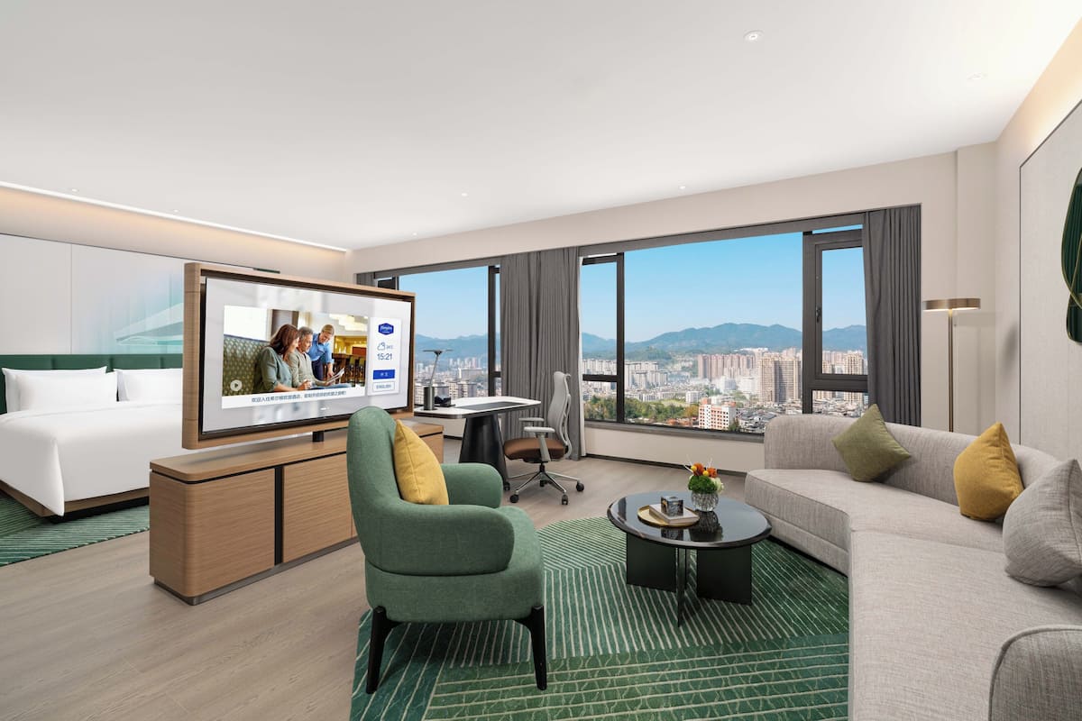 A modern hotel room with a large TV, a sofa, a green armchair, a desk, and a view of the cityscape.