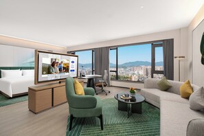 A modern hotel room with a large TV, a sofa, a green armchair, a desk, and a view of the cityscape.