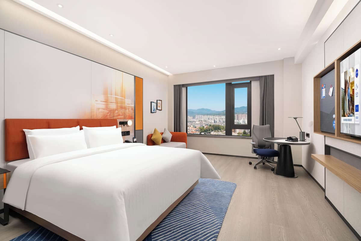 A hotel room with a large bed, a desk, a chair, a TV, and a window with a city view.