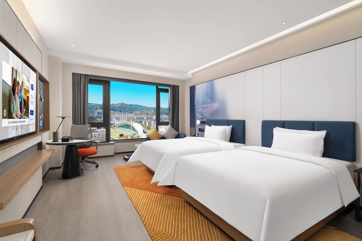 A hotel room with two beds, a desk, a TV, and a large window with a city view.