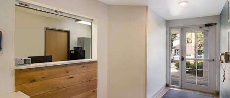 A hotel lobby with a wooden reception desk, a large mirror, a door with a glass panel, and a black mat at the entrance.