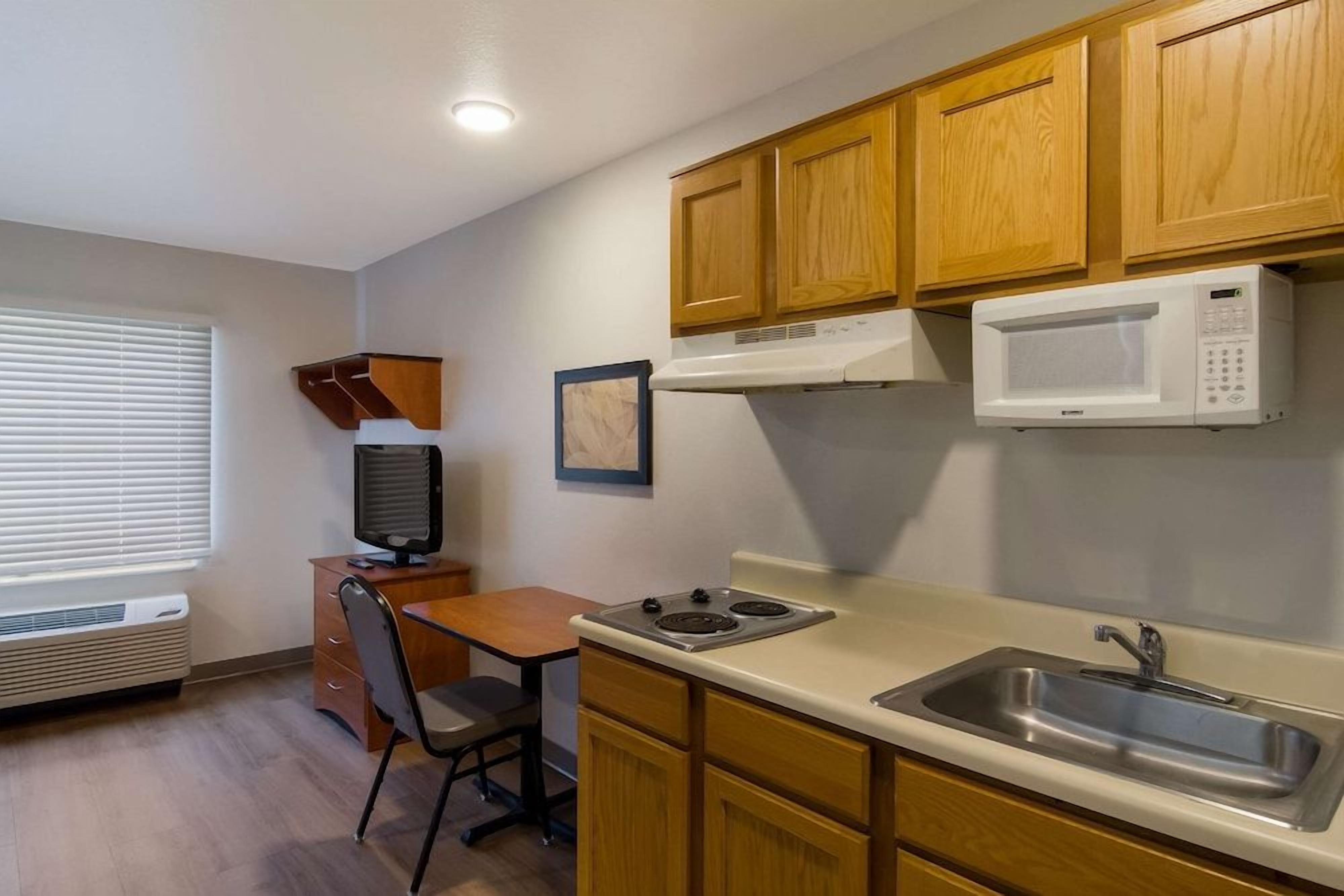 A compact kitchen with wooden cabinets, a microwave, a sink, a stove, a chair, and a small table.