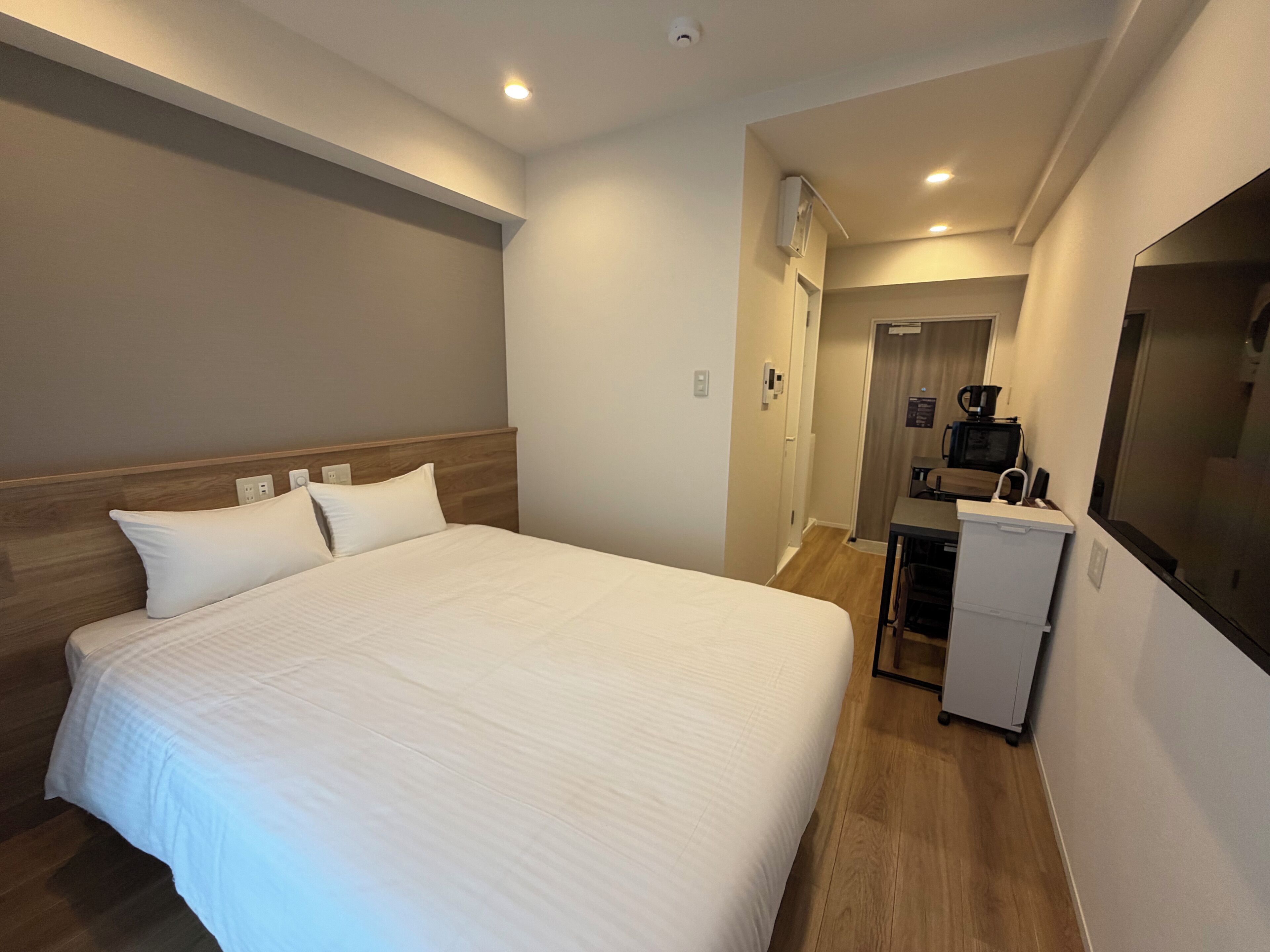 Quarto de hotel com uma cama grande, uma escrivaninha com computador e uma cozinha compacta.