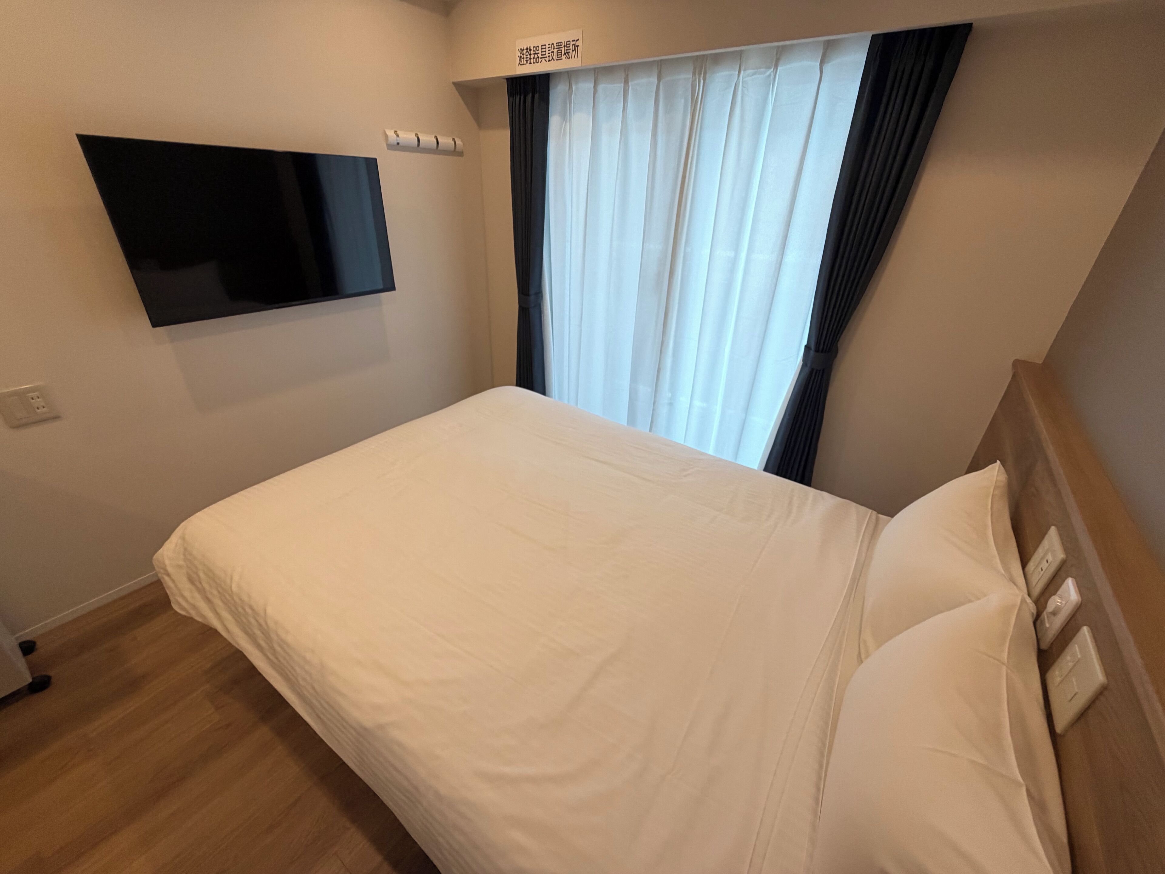 A hotel room with a bed, a flat-screen TV mounted on the wall, a window with curtains, and a wooden headboard.