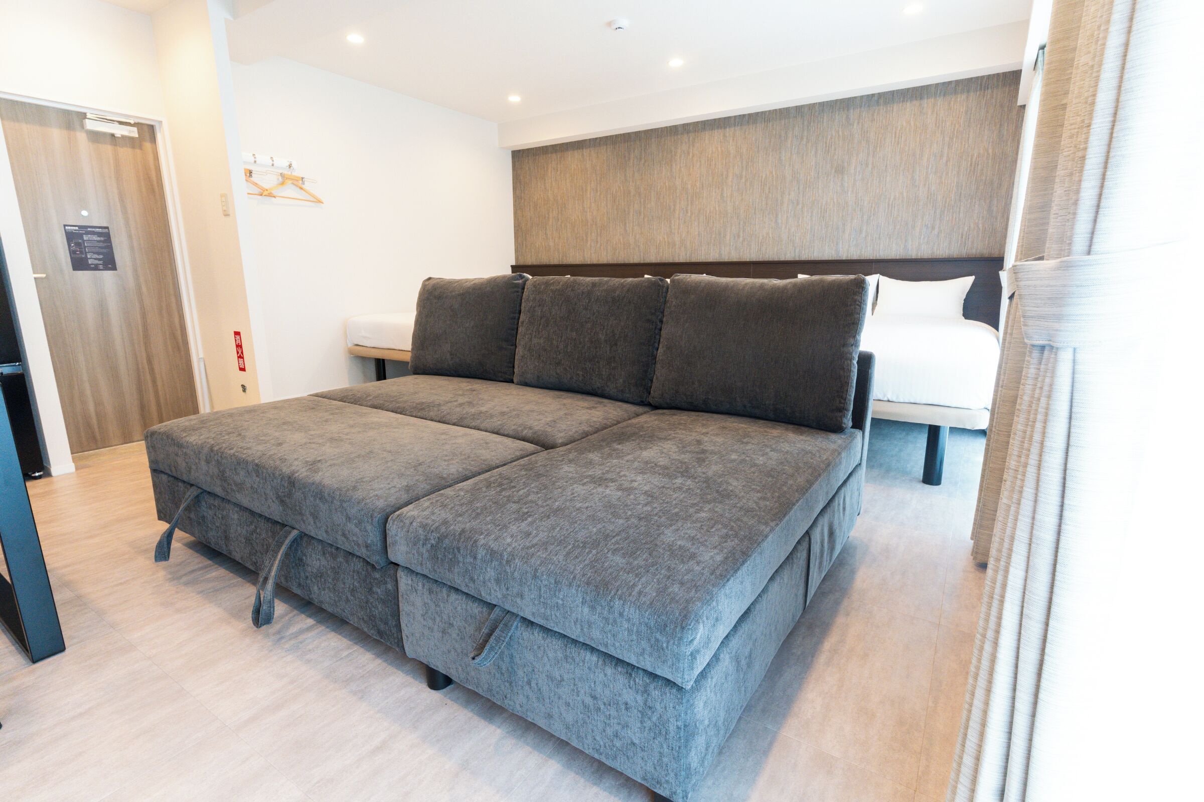 Quarto moderno com um sofá-cama cinzento, um painel de cama de madeira e uma parede branca.