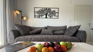 Un salon avec un canapé gris, un panier de fruits posé sur une table blanche et une œuvre d’art représentant un arbre accrochée au mur.